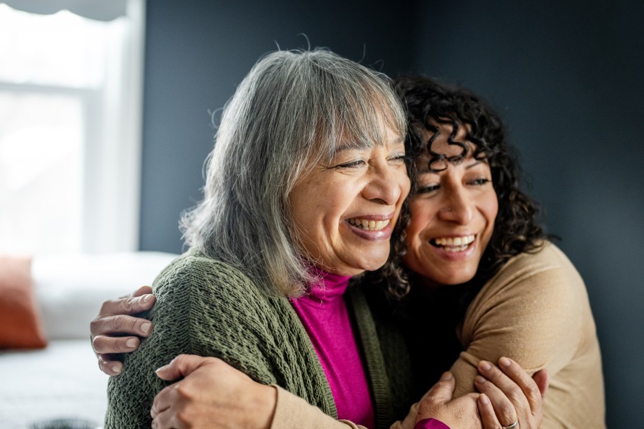 Caregiver hugging her senior mother at home, both smiling warmly and sharing a joyful, supportive moment.
