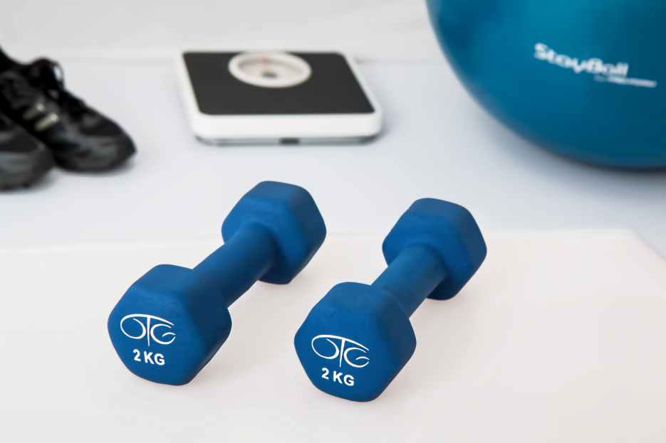 Light blue dumbbells on a workout mat with a scale, exercise ball, and shoes in the background, representing healthy routines for seniors.