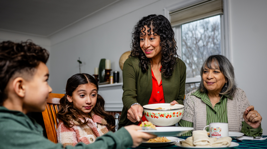 Family caregiver serving a meal to her children and senior mother at home, illustrating caregiving support across generations.
