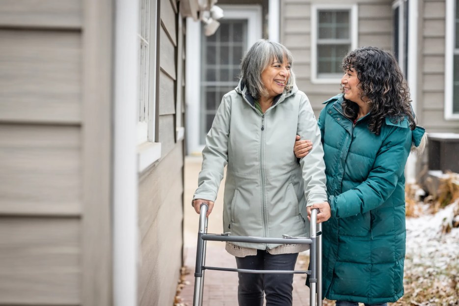 Family caregiver walking outside with her senior mother who uses a walker, sharing a supportive and caring moment together.