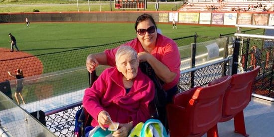 Caregiver assisting elderly woman in wheelchair with baseball field backdrop