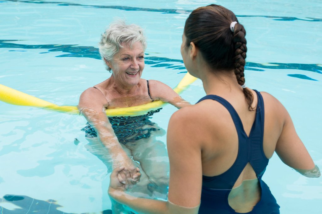 An elderly lady in a pool holding hands with an instructor, emphasizing the therapeutic nature of swimming for seniors