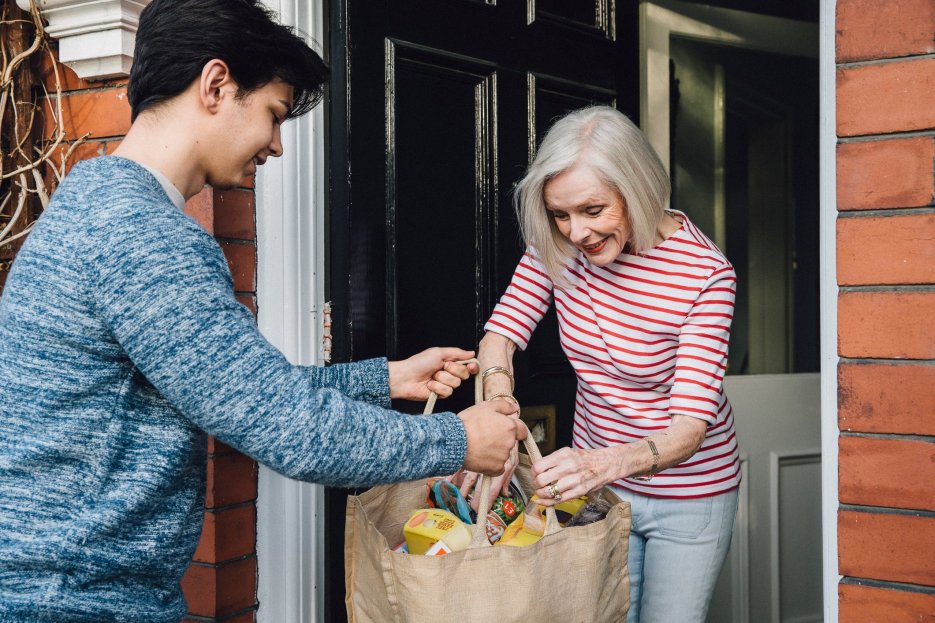 Caregiver delivering groceries to a senior woman at her doorstep, providing caregiving support and assistance with daily needs.