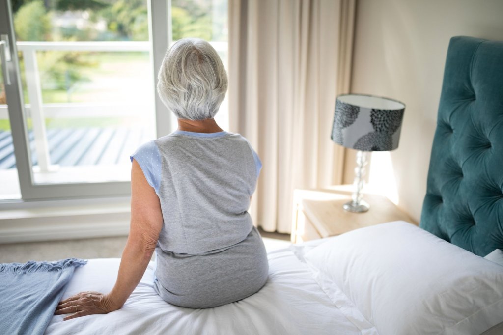 A caregiver sits on the edge of her bed, gazing out the window