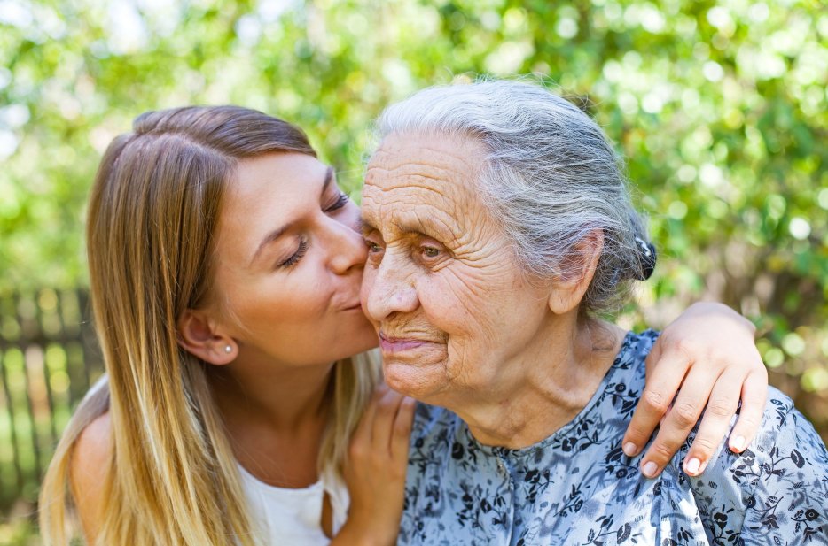 Family caregiver kissing her elderly mother on the cheek outdoors, showing love, compassion, and caregiving support.