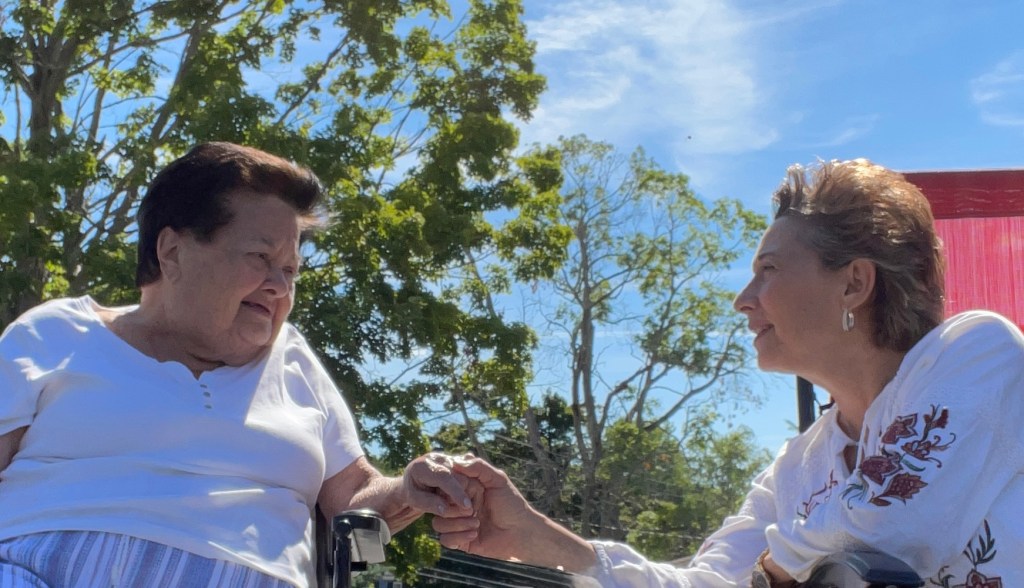 Linda gently holds her mother’s hand outdoors, both smiling warmly at each other. The moment reflects love, connection, and caregiving across generations.