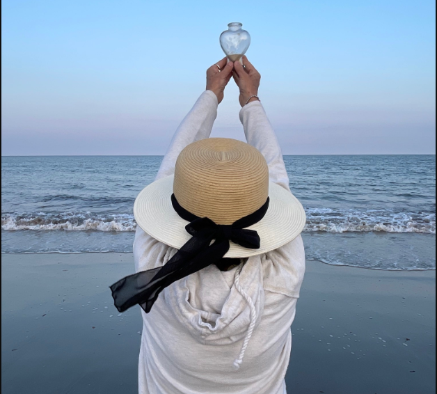 A caregiver stands on the shore at sunset, holding a heart-shaped urn above her head in a gesture of farewell.