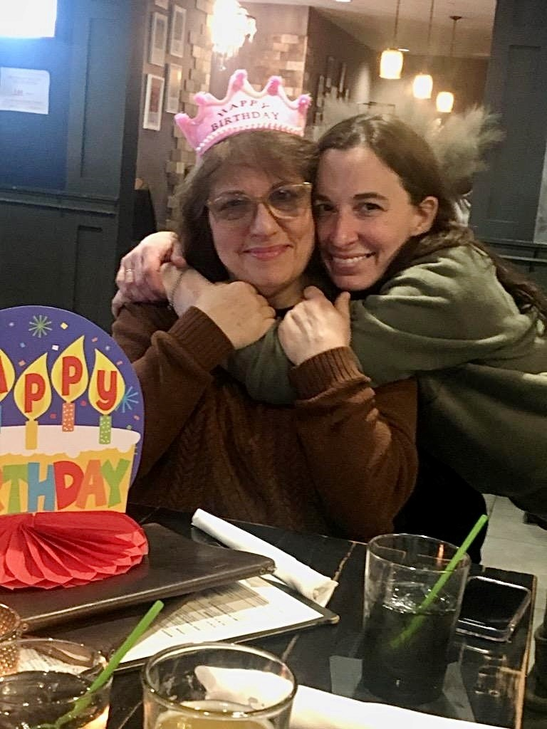 Two women hugging closely at a birthday celebration; the older woman wears a pink “Happy Birthday” crown while the younger woman embraces her from the side, both smiling warmly at the camera.
