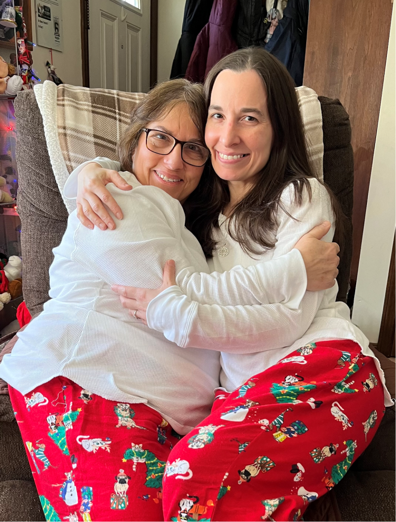A mother and her daughter, whom she cares for, sitting together in matching pajamas and hugging while smiling at the camera.