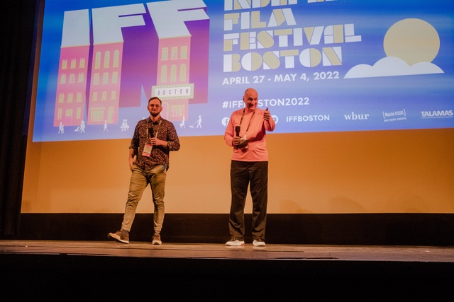 Two people stand on stage at the Independent Film Festival Boston, speaking in front of a large screen during a presentation about their caregiving documentary.