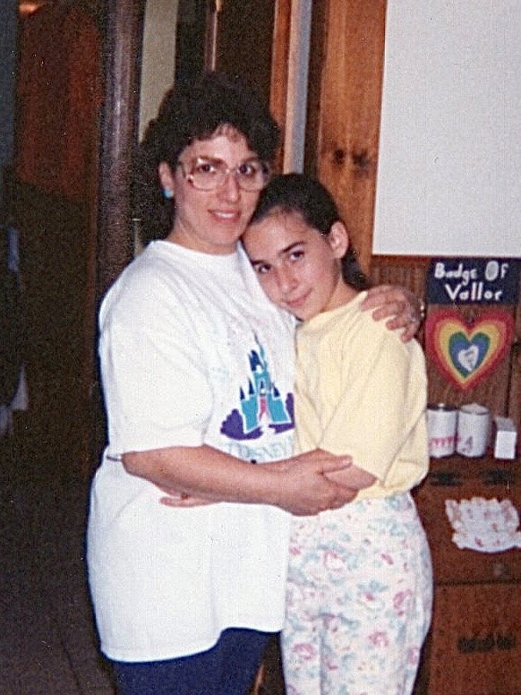 An older woman and a young girl standing together indoors, hugging closely; the mother has her arms around her daughter as they both look toward the camera, illustrating the family’s long medical journey and caregiving bond.